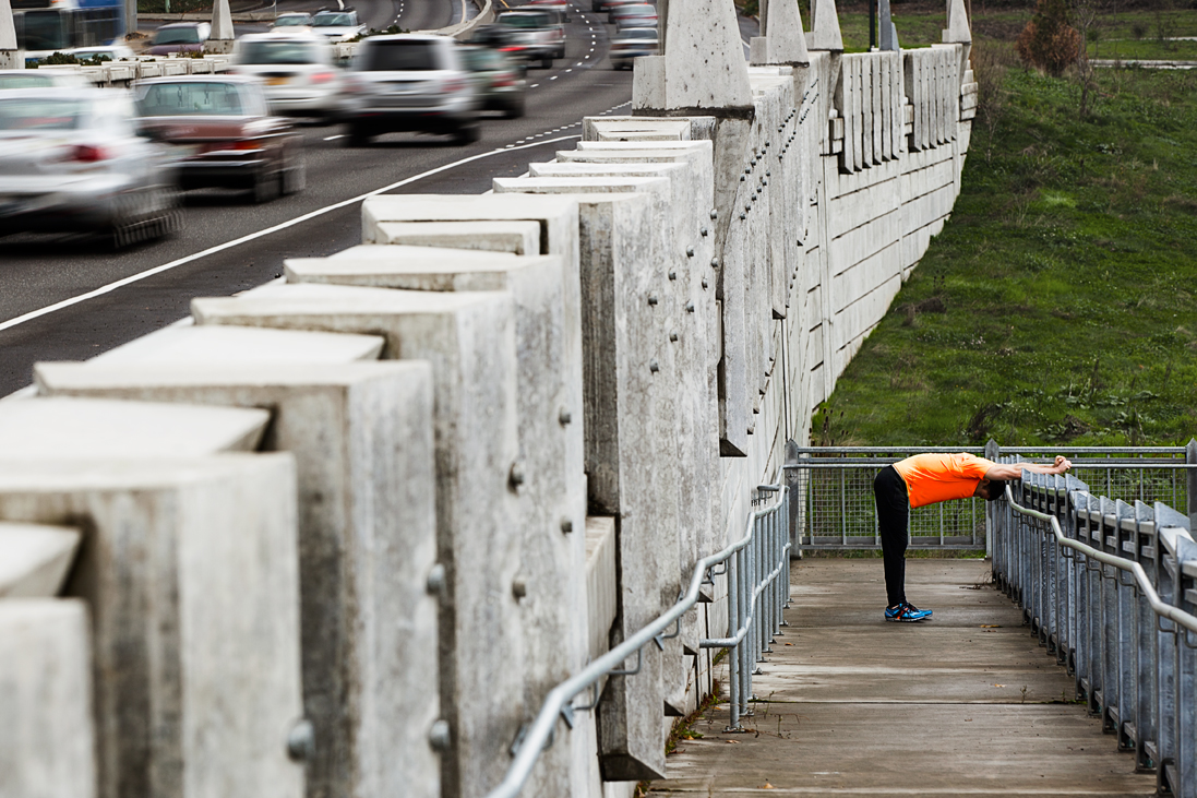 Runner in orange shirt stretching on ramp in urban city landscape, highway traffic