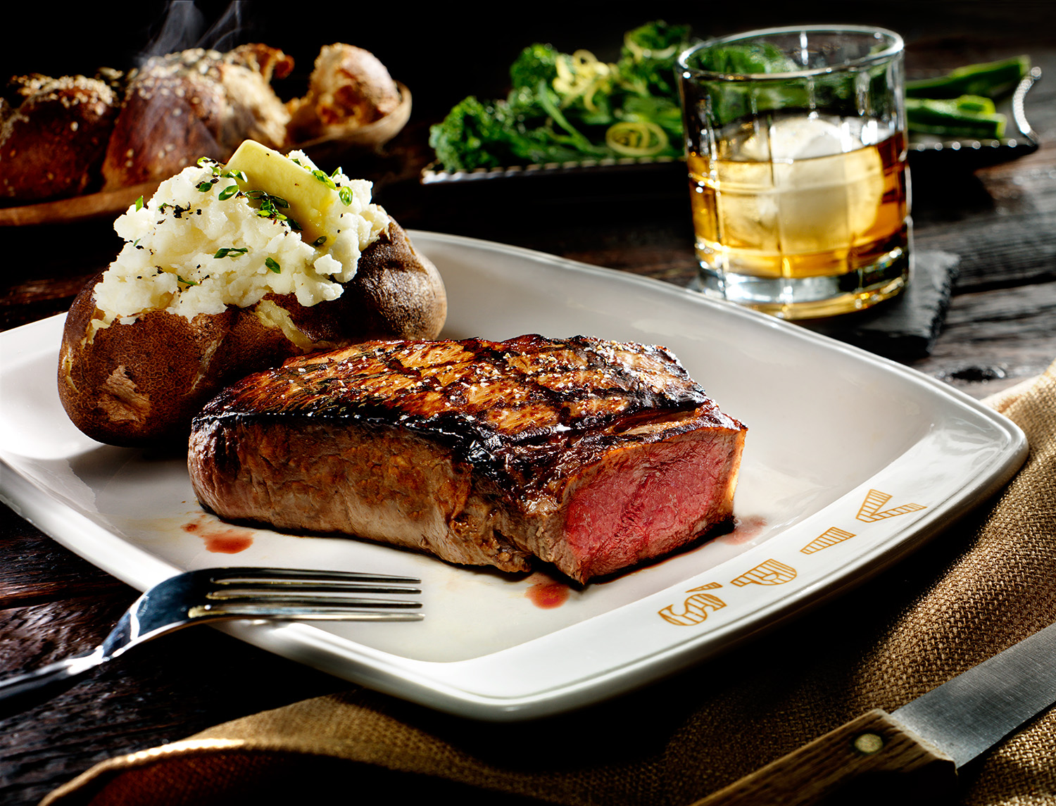Juicy steak and steaming baked potato steakhouse restaurant dinner on table with food dishes and drink